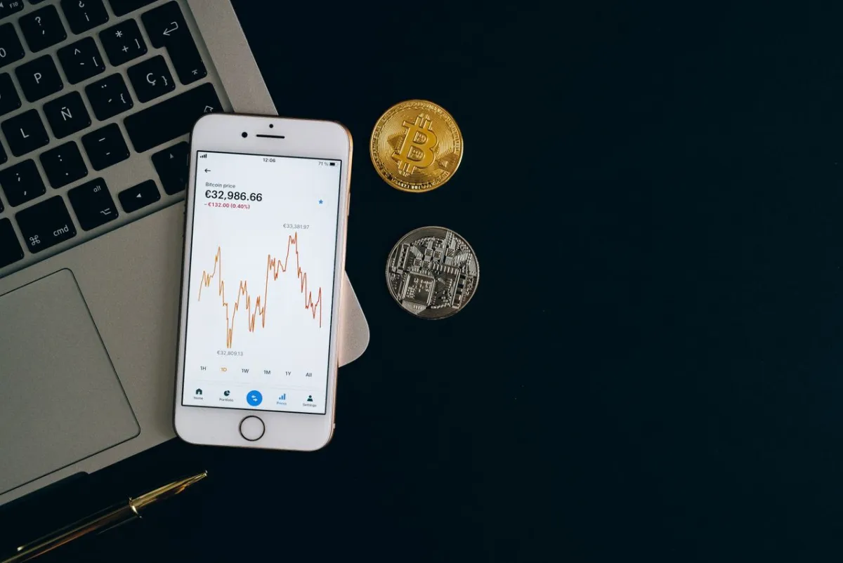 A phone showing a Bitcoin price chart beside two coins